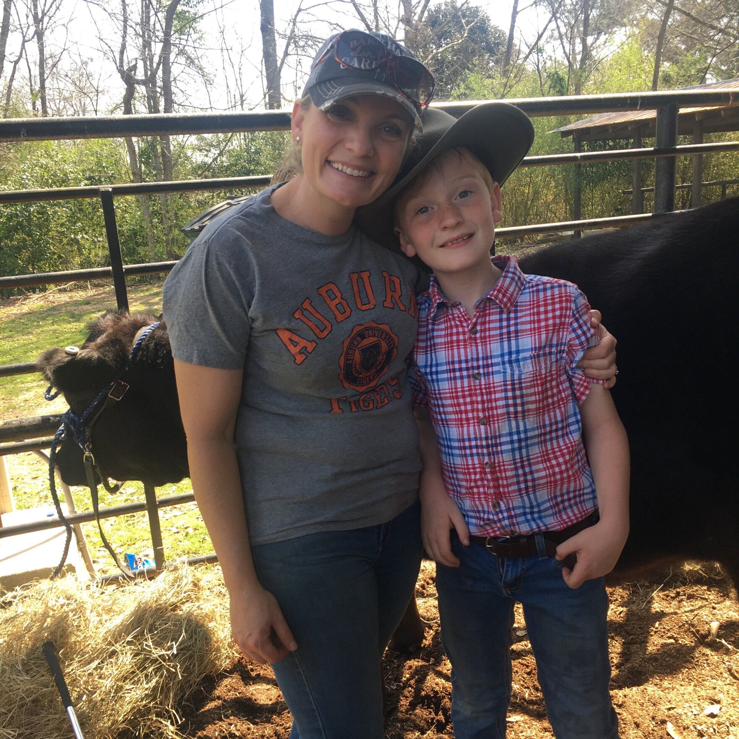 A woman and a young boy stand together smiling in a farm setting, with a black cow and hay in the background. The woman wears an Auburn Tigers t-shirt and a baseball cap, while the boy is dressed in a red and blue plaid shirt and jeans. This image captures a joyful moment of family bonding on a farm.
