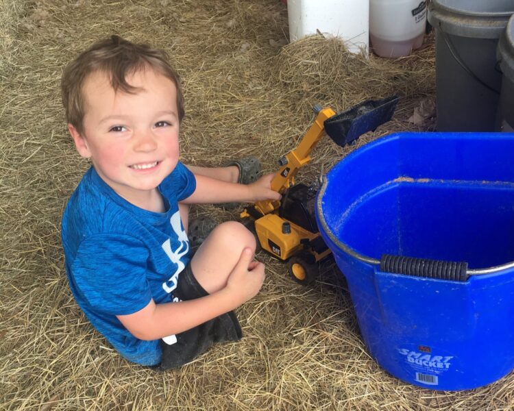 Smiling child playing with a yellow toy construction vehicle next to a blue bucket on straw-covered ground, showcasing outdoor play and imaginative activities.