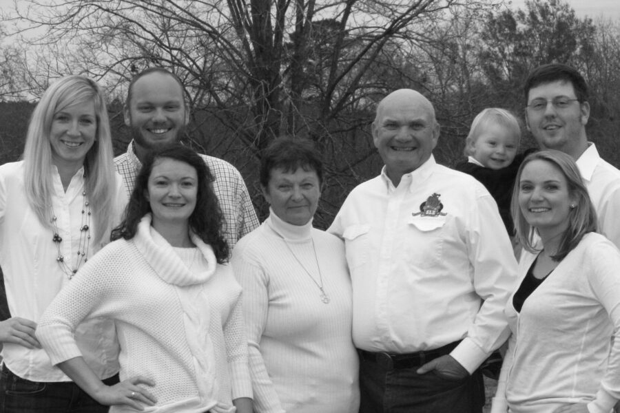 Black and white family portrait featuring eight members standing together outdoors, smiling. They are dressed in casual white and light-colored clothing, with a backdrop of bare trees and a cloudy sky, conveying a warm and friendly family gathering.
